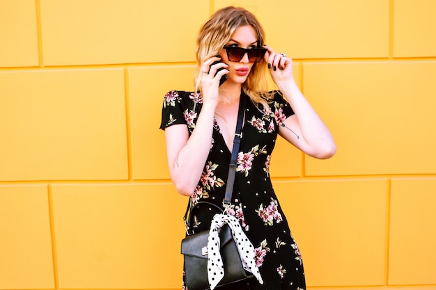 Elegante modieuze vrouw poseren in de buurt van gele muur en spreken door haar smartphone