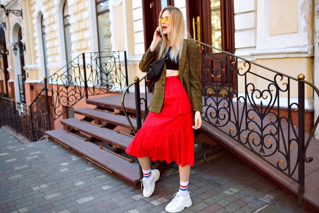 Elegante trendy blonde vrouw die zich voordeed op straat in de buurt van prachtig oud gebouw, sprekend door haar telefoon, modieuze trendy hipster outfit en zonnebril dragen, lente herfst stijl.
