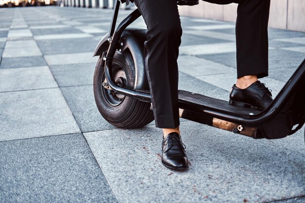 Elegante zakenman in zwart pak zit op zijn elektrische scooter in de buurt van zijn kantoor.