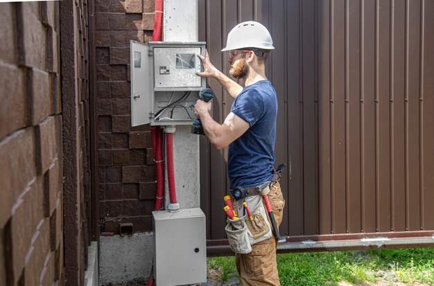 Elektricien-bouwer op het werk, onderzoekt de kabelverbinding in de elektrische leiding in de romp van een industrieel schakelbord. Professioneel in overall met gereedschap voor elektriciens.