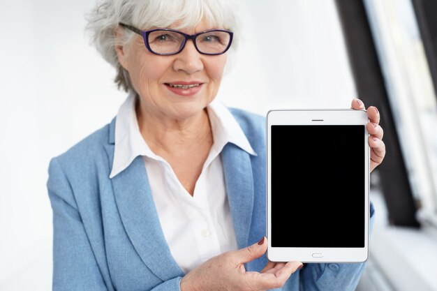 Elektronische apparaten, gadgets, technologie en verbindingsconcept. Vrolijke elegante oudere Europese grijze haren vrouw in brillen presenteren digitale tablet met zwart scherm met copyspace voor uw tekst