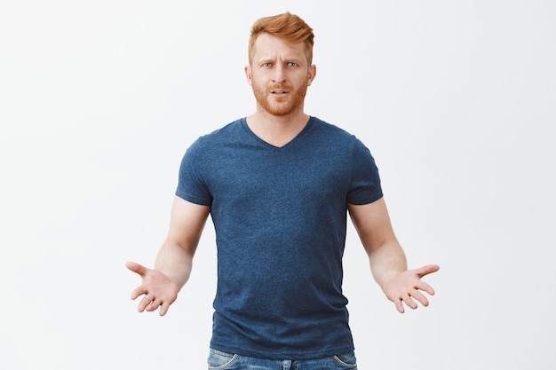 Elk probleem. Portret van verwarde ondervraagde aantrekkelijke mannelijke Europese roodharige man in blauw t-shirt, gebarend met handpalmen in clueless pose, wachtend op uitleg over grijze muur