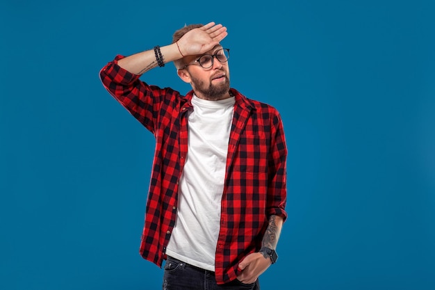 Emotioneel en mensenconcept: jonge, bebaarde man in geruit overhemd. Hipster-stijl. Studio opname op blauwe achtergrond. Ruimte kopiëren