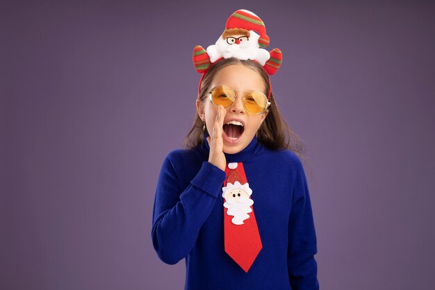 Emotioneel meisje in blauwe coltrui met rode stropdas en grappige kerstrand op het hoofd schreeuwend met de hand in de buurt van de mond die over de paarse muur staat