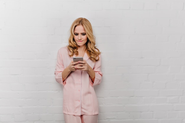 Emotioneel meisje in katoenen pyjama's poseren met telefoon op witte muur. Mooie gekrulde vrouw in roze nachtkostuum smartphonescherm kijken.