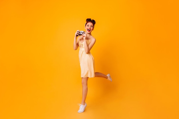 Emotioneel pinup meisje met camera dansen op gele achtergrond Studio shot van vrouwelijke fotograaf in jurk
