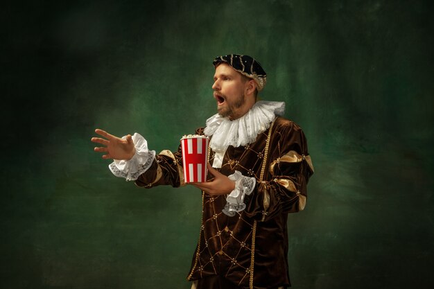 Emotioneel voetbal kijken. Portret van middeleeuwse jonge man in vintage kleding staande op een donkere achtergrond. Mannelijk model als hertog, prins, koninklijk persoon. Concept vergelijking van moderne tijdperken, mode.