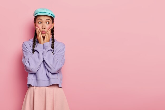 Emotionele Aziatische vrouw met verbluffende look, vergeet iets belangrijks, kijkt weg op vrije ruimte, gekleed in stijlvolle kleding, poseert over roze muur