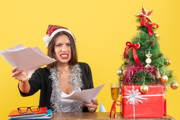 Emotionele boze zakelijke dame in pak met kerstman hoed en nieuwjaarsversieringen met documenten en zittend aan een tafel met een kerstboom