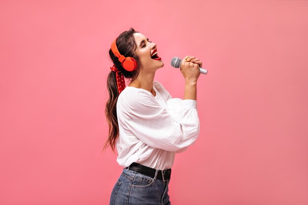 Emotionele dame in rode koptelefoon zingen in de microfoon. Helder mooi meisje in witte blouse en in spijkerbroek met zwarte gordel poseren.