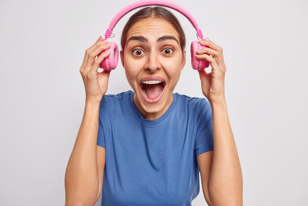 Emotionele donkerharige jonge Europese vrouw zet koptelefoon af met luide muziek roept luid houdt mond open, gekleed in casual blauw t-shirt geïsoleerd op witte achtergrond Wat zei je