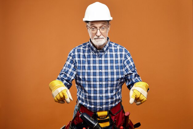 Emotionele grappige bejaarde senior huis bouwer dragen van beschermende helm, veiligheidshandschoenen en tas met instrumenten rond zijn middel hebben geschokt verrast gelaatsuitdrukking, met de voorste vingers naar beneden gericht