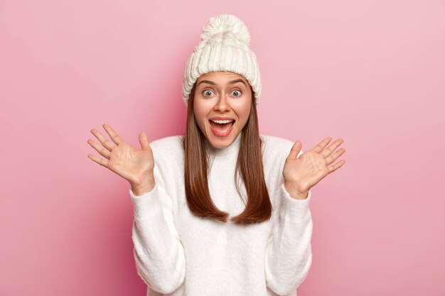 Emotionele jonge blanke vrouw werpt handpalmen, houdt de mond open, heeft plezier, gekleed in witte winterkleren, vormt op roze achtergrond.