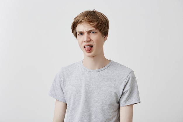 Emotionele jonge europese man met lichtgrijs t-shirt grijnzend, monden makend, zijn tong uitstekend om iemand te plagen, gedraagt zich als een klein stout kind