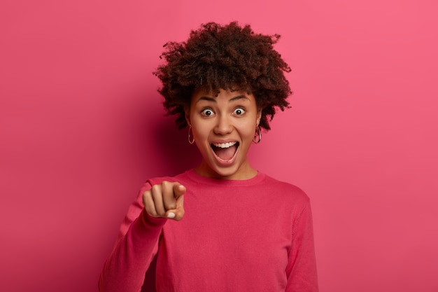 Emotionele jonge vrouw met een donkere huid ziet er opgewonden en opgewekt uit, wijst direct met de wijsvinger, kiest iets of kiest iemand, draagt een roze trui, poseert binnen. mensen en keuze.