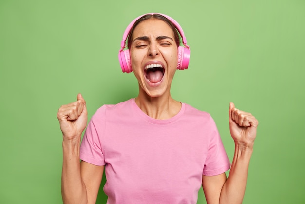 Emotionele jonge vrouw schreeuwt van opwinding balt vuisten houdt mond wijd open schreeuwt van succes luistert muziek in koptelefoon draagt roze t-shirt geïsoleerd over levendige groene muur
