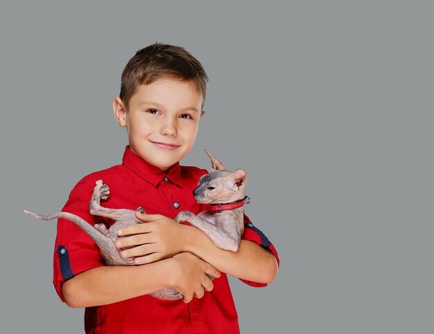 Emotionele kleine jongen in een rood polot-shirt houdt een kat vast. Geïsoleerd op grijze achtergrond.