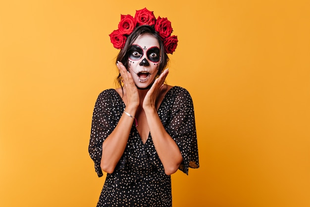 Emotionele Mexicaanse donkerharige vrouw met bloemen op haar hoofd maakt geschokt gezicht zichzelf aan te raken met haar handen