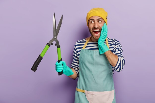 Emotionele onderhoudsmedewerker houdt snoeischaar, gekleed in werkkleding, snijdt takken, geïsoleerd op violette achtergrond.