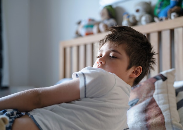 Emotionele portret jonge jongen liggend op bed, slaperig kind 's ochtends wakker in zijn slaapkamer met ochtendlicht, kind van 7 jaar oud ontspannen in de slaapkamer. gezondheidszorg voor kinderen of slaapproblemen bij jong kind