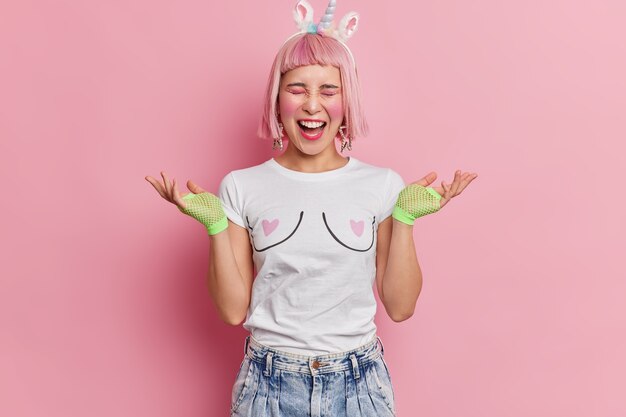 Emotionele rozeharige jonge Aziatische vrouw spreidt handpalmen uit en roept luid gekleed in een casual t-shirt en jeans heeft lichte make-up