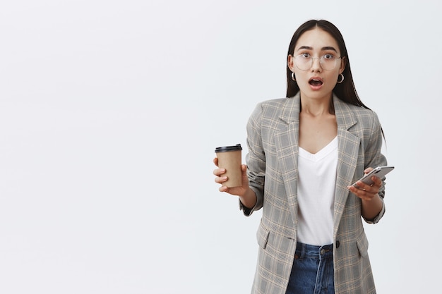 Emotionele stijlvolle vrouwelijke student in jas en bril, smartphone en kopje koffie te houden, praten met verbazing