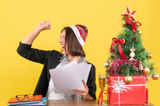 Emotionele trotse zakelijke dame in pak met kerstman hoed en nieuwjaarsversieringen met documenten en zittend aan een tafel met een kerstboom erop in het kantoor