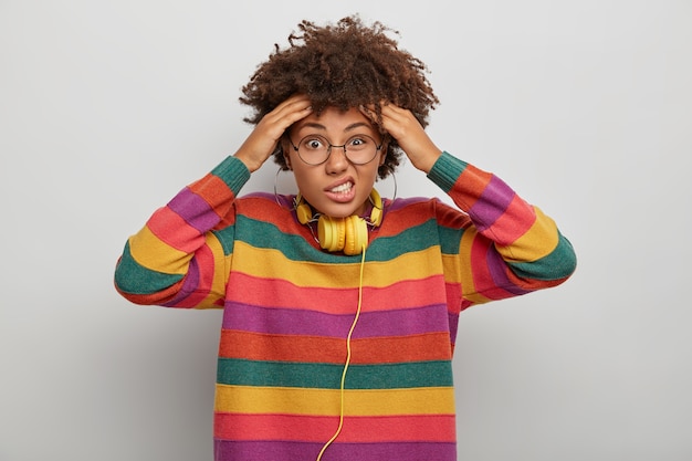 Emotionele verbaasd jonge Afro-Amerikaanse dame houdt beide handen op het hoofd, fronst gezicht en kijkt met ongenoegen, lijdt aan hoofdpijn