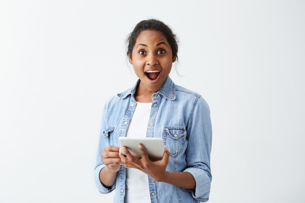 Emotionele verbaasde jonge vrouw met een donkere huidskleur en een blauw shirt met een tablet in haar handen, verrast en geschokt door positief onverwacht nieuws. Menselijke gezichtsuitdrukkingen en emoties