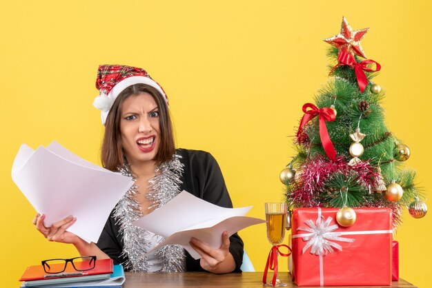 Emotionele verward zakelijke dame in pak met kerstman hoed en nieuwjaarsversieringen met documenten en zittend aan een tafel met een kerstboom erop in het kantoor