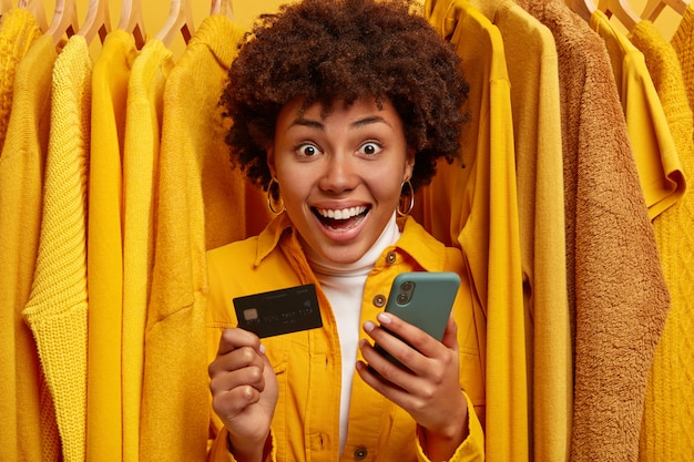 Emotionele vrolijke winkelende vrouw gebruikt mobiele telefoon om online te betalen, houdt creditcard vast, staat tussen gele truien op hangers