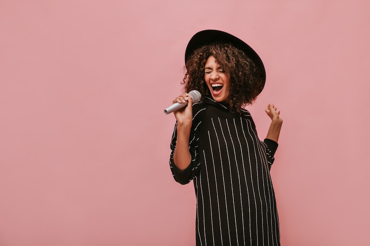 Gratis Foto emotionele vrouw met krullend donkerbruin kapsel in stijlvolle hoed en gestreepte zwarte jurk met microfoon en zingen op roze muur..