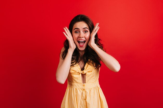 Emotionele vrouw schreeuwt gelukkig op rode muur. Krullende dame in geel geruite outfit poseren.