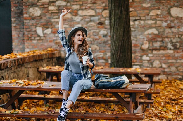 Emotionele vrouwelijke model in korte jeans en zwarte schoenen drinkt koffie in park in warme ochtend van september