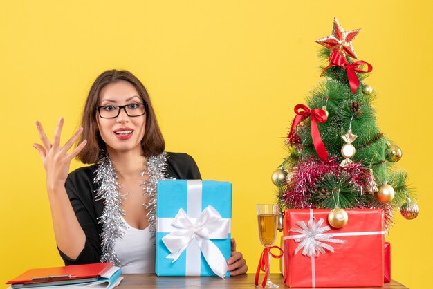 Emotionele zakelijke dame in pak met bril met haar cadeau en zittend aan een tafel met een kerstboom erop in het kantoor