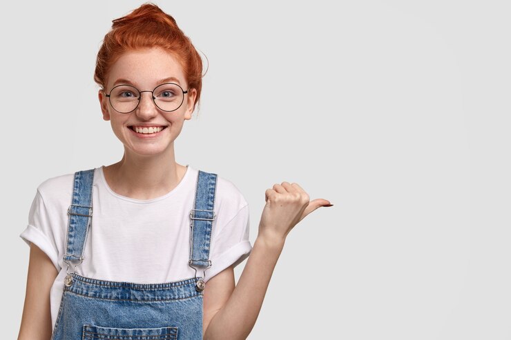 Gratis Foto energieke tevreden tienermeisje met roodgekamd haar in een knot, draagt een bril, een tuinbroek in spijkerbroek, wijst opzij naar de vrije ruimte, demonstreert iets plezierigs, wijst de weg. mensen, reclame