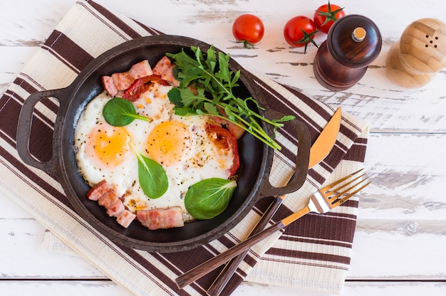 Engels ontbijt of lunch met gebakken ei, spinazie, rucola, tomaten en spek op zwarte pan.