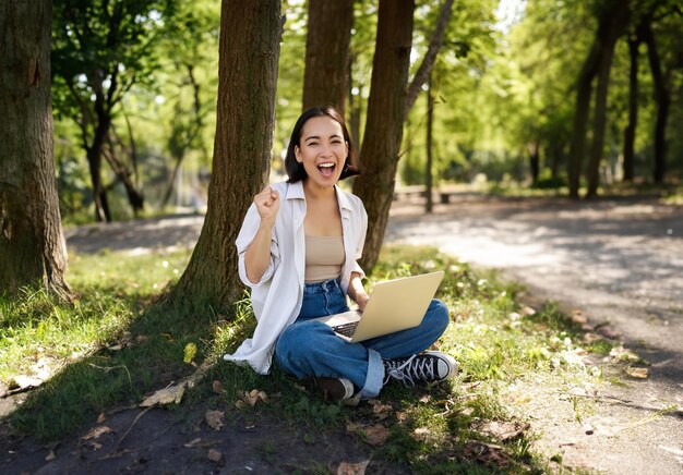 Enthousiast jong Aziatisch meisje dat met laptop naast de boom zit in een groen zonnig park en triomf viert