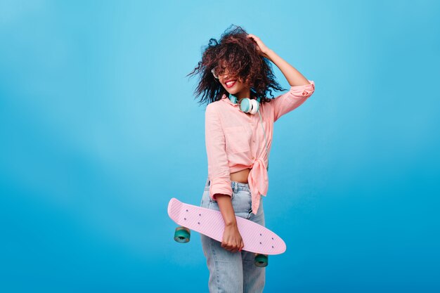 Enthousiast meisje met bruin krullend kapsel genieten. Slank Afrikaans meisje met skateboard spelen met haar haren en lachen.