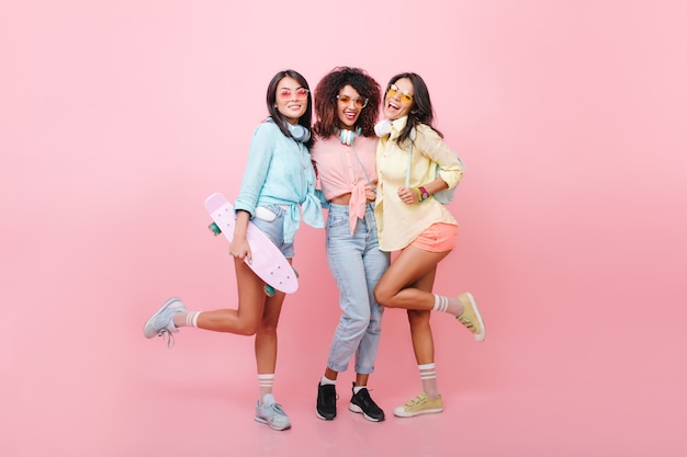 Enthousiast zwartharig vrouwelijk model met mooie tan die zich op één been bevindt terwijl haar vrienden glimlachen. Vrolijke Afrikaanse vrouw in jeans en zwarte schoenen poseren in de buurt van skater meisje in blauw shirt.