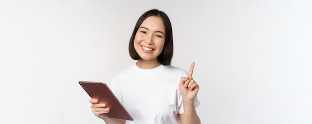 Enthousiaste aziatische vrouw met een tablet die de vinger opsteekt en er verbaasd uitziet terwijl ze naar boven wijst