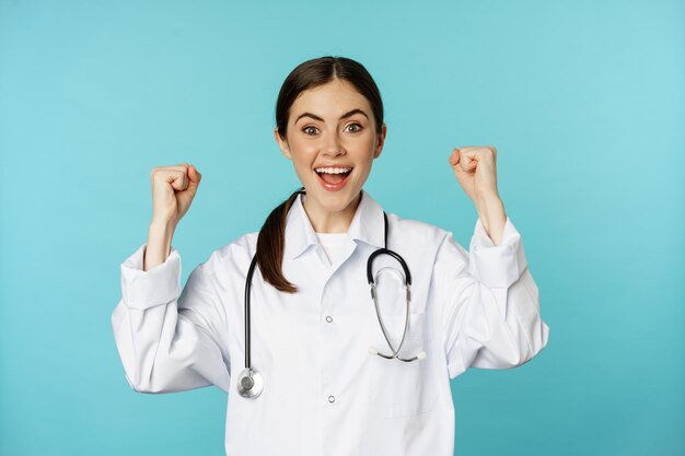 Enthousiaste doktersvrouw, zich verheugend, winnend en vierend, doel bereiken, in witte jas tegen blauwe achtergrond staan
