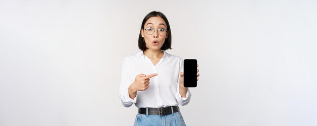 Enthousiaste jonge aziatische vrouw die met de vinger wijst naar het smartphonescherm met advertenties op de witte achtergrond van de mobiele telefoon