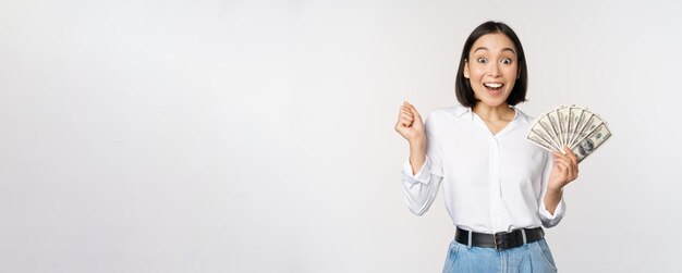 Enthousiaste jonge aziatische vrouw die opgewonden kijkt naar de camera die gelddollars in de hand houdt en over een witte achtergrond staat