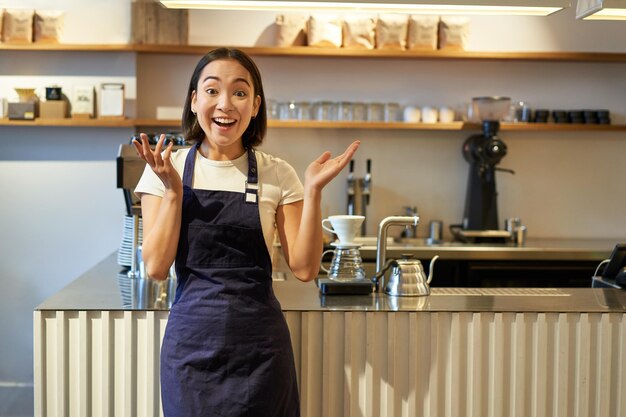 Enthousiaste jonge aziatische vrouwenbarista die in schort verbaasd kijkt klapt handen en glimlacht status in co