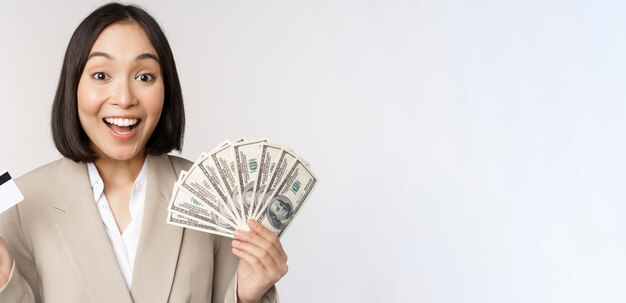 Enthousiaste jonge Aziatische zakenvrouw die gelddollars en creditcard toont die zich over witte achtergrond bevinden