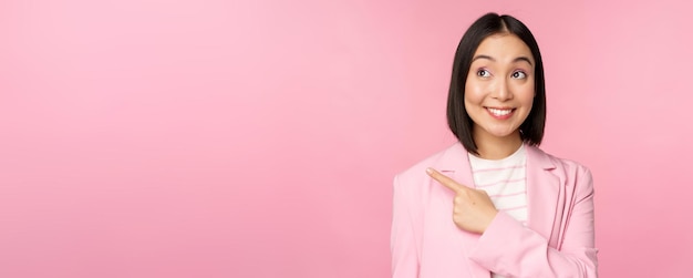 Enthousiaste jonge Aziatische zakenvrouw kantoormedewerker wijzende vinger naar links kijkend naar banneradvertentie met een gelukkige glimlach met advertentie roze achtergrond