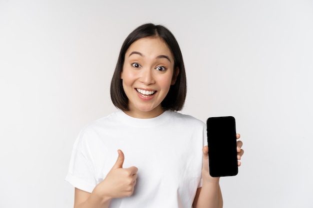 Enthousiaste jonge vrouw die duim omhoog laat zien en het scherm van de mobiele telefoon in een t-shirt op een witte achtergrond