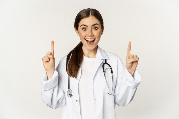 Enthousiaste jonge vrouwelijke arts lacht, wijst met de vingers omhoog, draagt ziekenhuisuniform, staat op een witte achtergrond