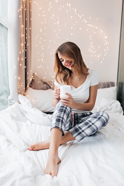 Enthousiaste op blote voeten blanke meisje poseren in bed. Blij dat jonge vrouw in geruite broek plezier heeft in de ochtend.